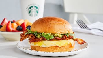 A breakfast sandwich on a plate with a Starbucks cup in the background.