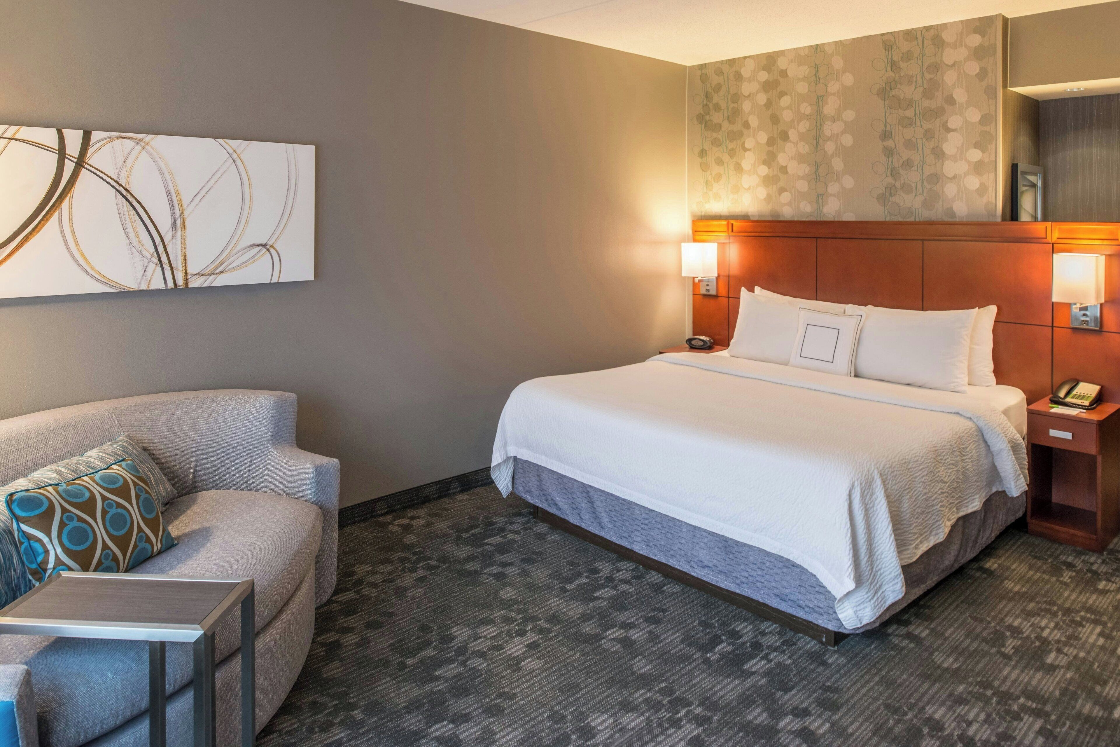 A hotel room with a bed, a sofa, a small table, and a wall-mounted artwork.