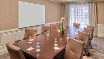 A conference room with a large table, eight chairs, a projector screen, and a potted plant.