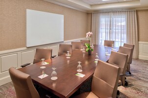 Un salón de conferencias con una gran mesa, ocho sillas, una pantalla de proyector y una planta en maceta.
