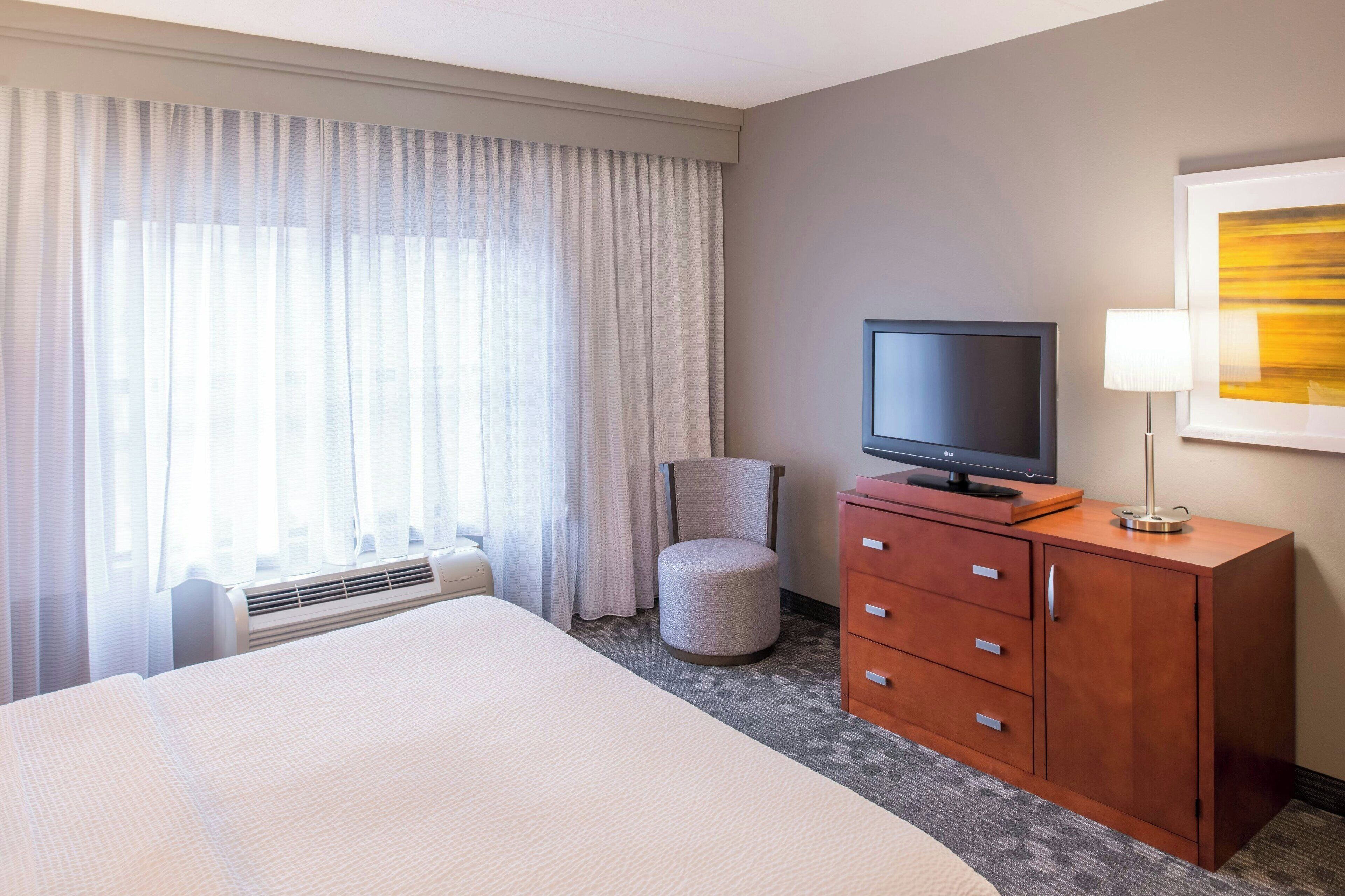 Une chambre d’hôtel avec un lit, un téléviseur posé sur une commode en bois, une chaise et une grande fenêtre avec des rideaux.