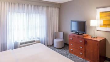 Quarto de hotel com cama, televisão sobre um móvel de madeira, uma cadeira e uma janela grande com cortinas.
