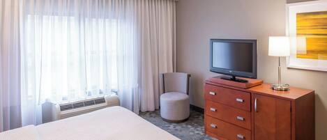 Une chambre d’hôtel avec un lit, un téléviseur posé sur une commode en bois, une chaise et une grande fenêtre avec des rideaux.