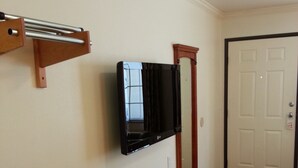 Habitación de hotel con televisor de pantalla plana montado en la pared, estante de madera, microondas y puerta con picaporte.