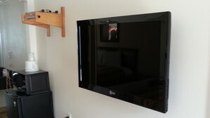 Una habitación con una televisión de pantalla plana montada en la pared, un pequeño estante de madera encima y un refrigerador negro a la izquierda.