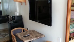 Una habitación pequeña con un televisor montado en la pared, dos sillas de madera con cojines, una mesita y un estante de madera sobre el televisor.