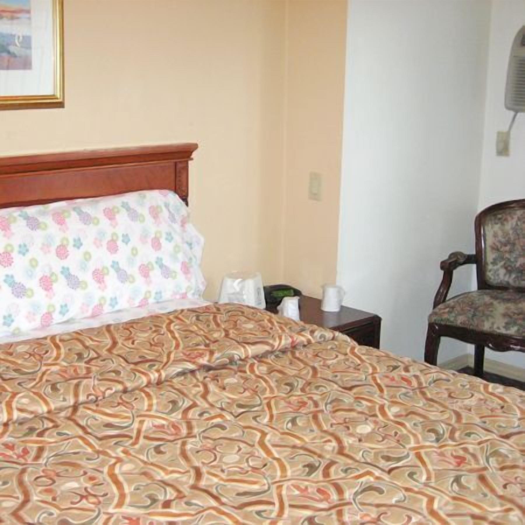 A hotel room with a bed, a chair, a nightstand, and a wall-mounted air conditioner.