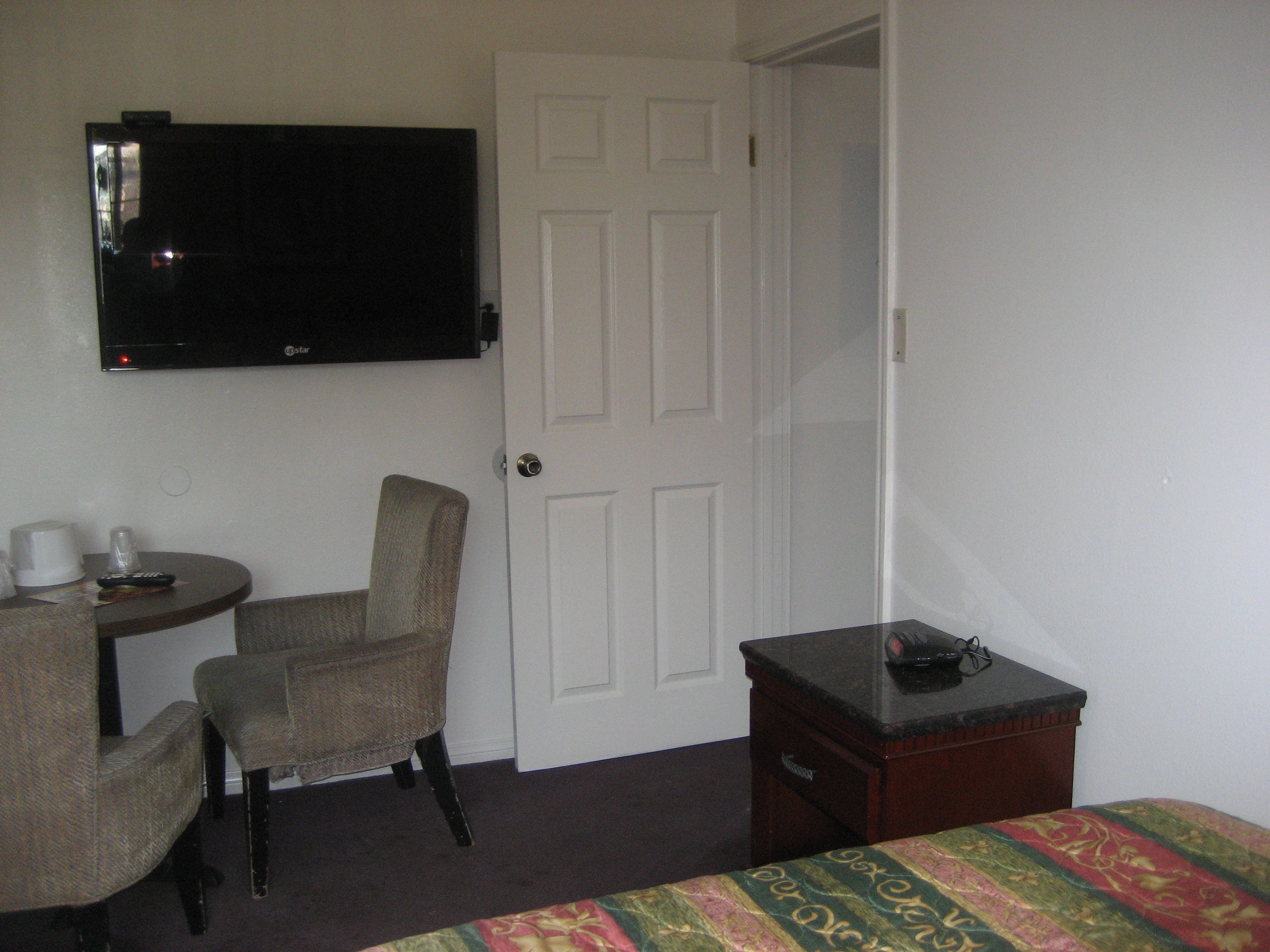 Una habitación de hotel con cama, escritorio con silla, televisión instalada en la pared y una puerta.