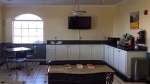 A kitchen with white cabinets, a black countertop, a dining table with chairs, a TV mounted on the wall, and a window with blinds. - Motel 6 Tyler, TX (Tyler)