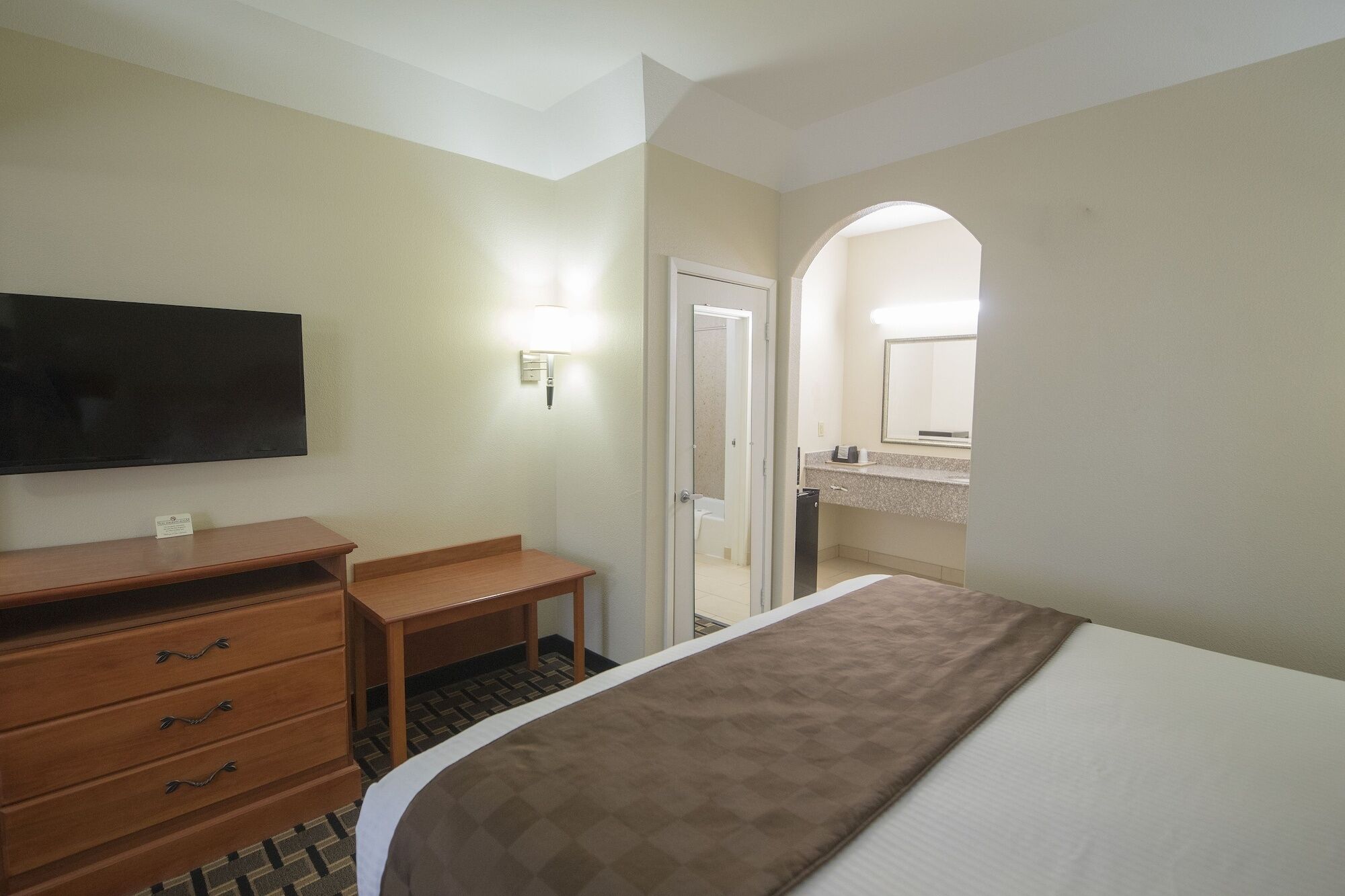 Une chambre d’hôtel avec un lit, une commode en bois, un téléviseur, un bureau et une salle de bain visible à travers une porte en arc de cercle.