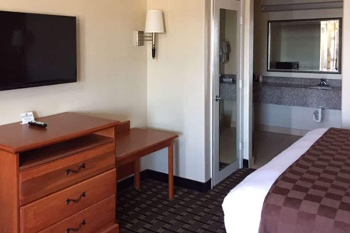 A hotel room with a flat-screen TV mounted on the wall, a wooden desk with drawers, a bedside table, and a checkered bedspread.