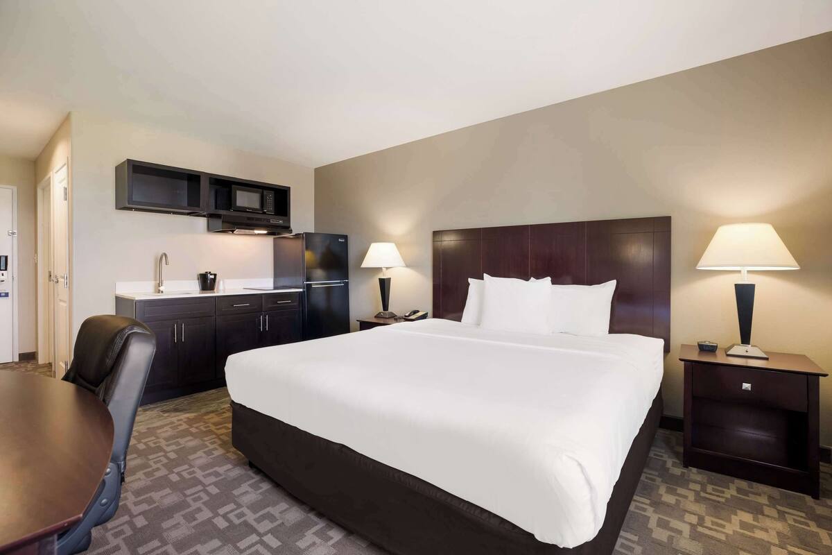 Een hotelkamer met een groot bed, een bureau, een stoel, een kleine kitchenette en twee bedlampen.