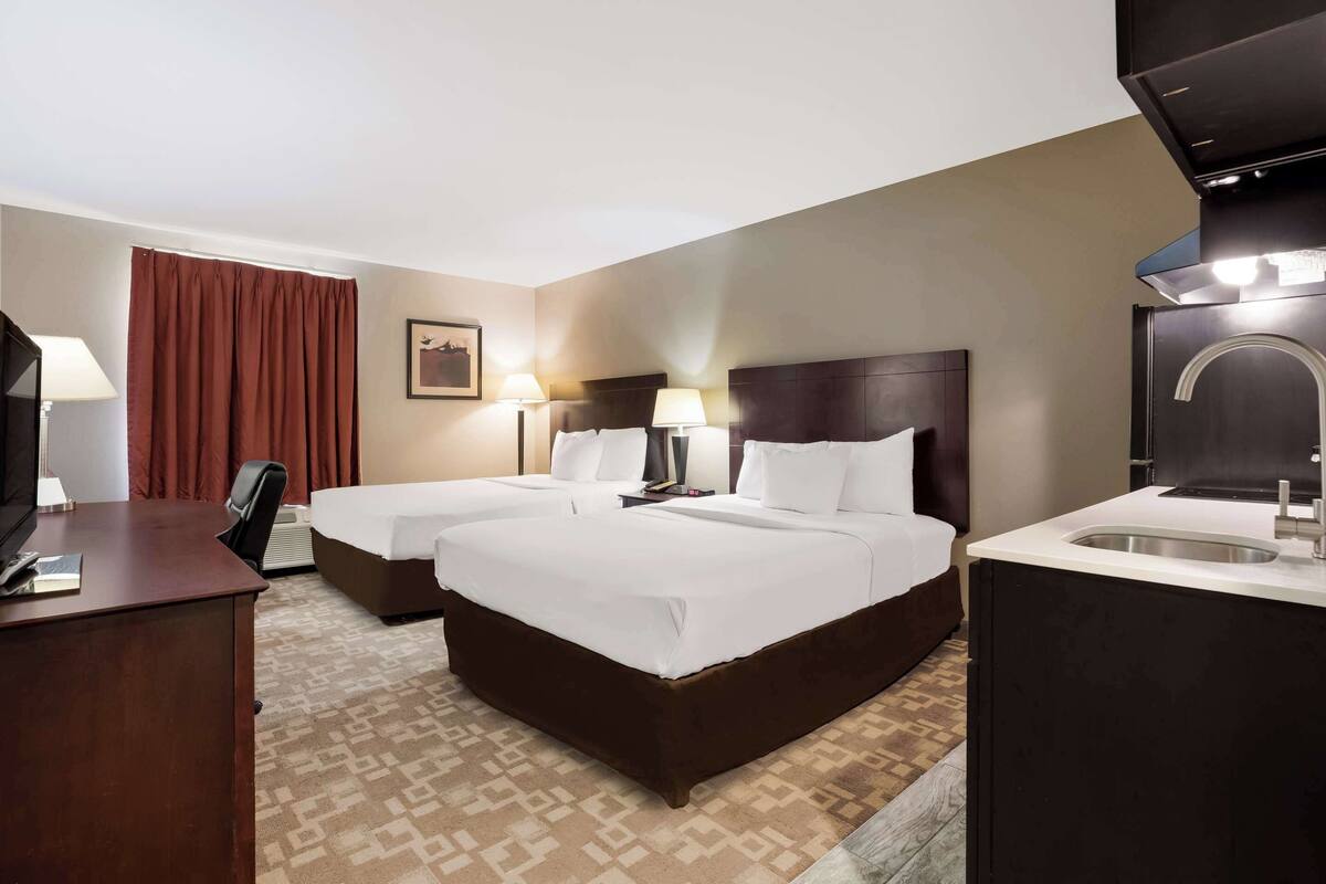 Hotelkamer met twee bedden, een bureau, een stoel, bureaulamp, een televisie aan de muur, een raam met gordijnen en een badkamer met wastafel en spiegel.