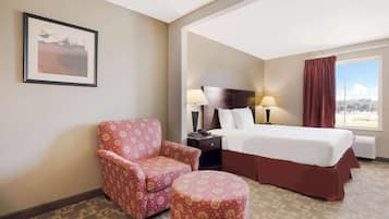 A hotel room with a bed, a chair, a small ottoman, a nightstand, a lamp, and a window with a view.