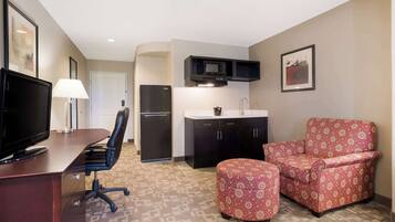 A hotel room with a TV, desk, office chair, kitchenette, and patterned armchair.