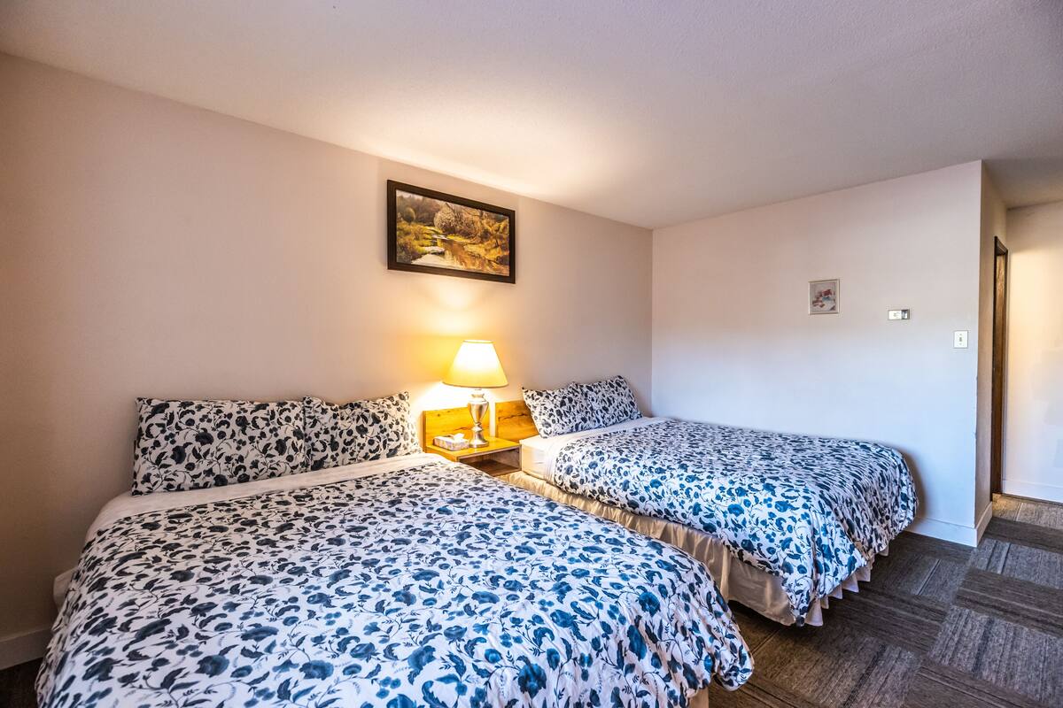A hotel room with two beds, a nightstand with a lamp, a framed picture on the wall, and a wooden floor.