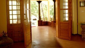 Un pasillo de madera con una puerta grande de madera a la derecha, una mesita con un florero y vistas al exterior con vegetación.
