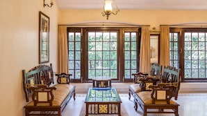A spacious room with a large window, a wooden sofa set, a glass-top coffee table, and a chandelier. - WelcomHeritage Golf View (Pipariya)