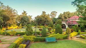 A well-maintained garden with a variety of shrubs and trees, a bench, and a building with a red roof. - WelcomHeritage Golf View (Pipariya)