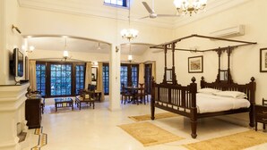 A spacious room with a four-poster bed, a dining area with a table and chairs, and large windows with a view of trees. - WelcomHeritage Golf View (Pipariya)