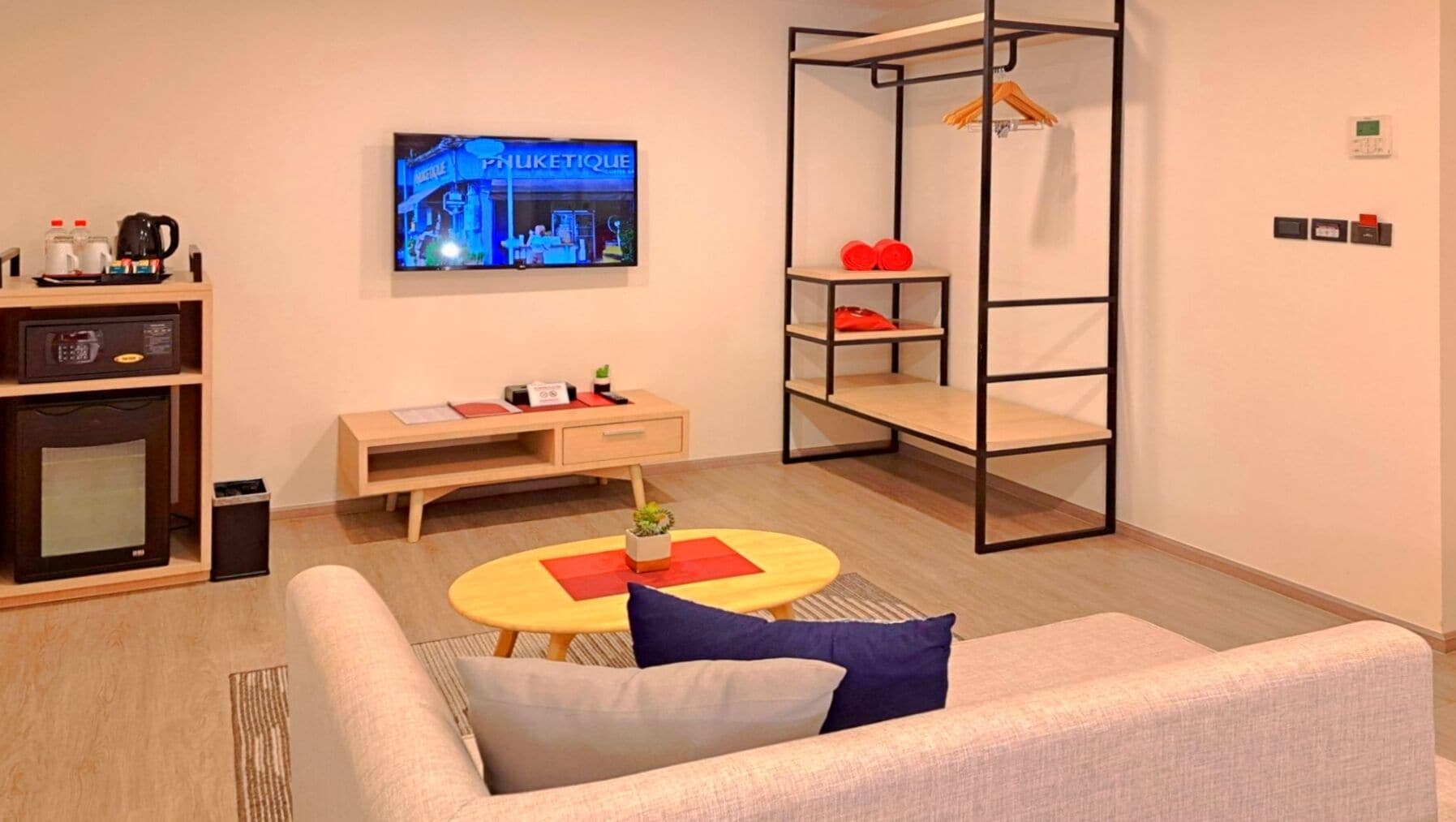 A modern living room with a sofa, a coffee table, a TV displaying a cityscape, a small desk with a kettle, and a metal shelf unit.