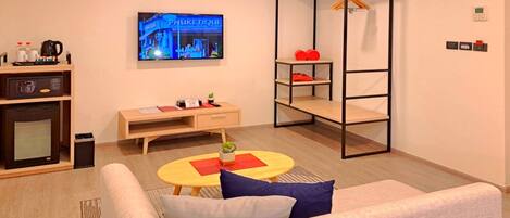 A modern living room with a sofa, a coffee table, a TV displaying a cityscape, a small desk with a kettle, and a metal shelf unit.