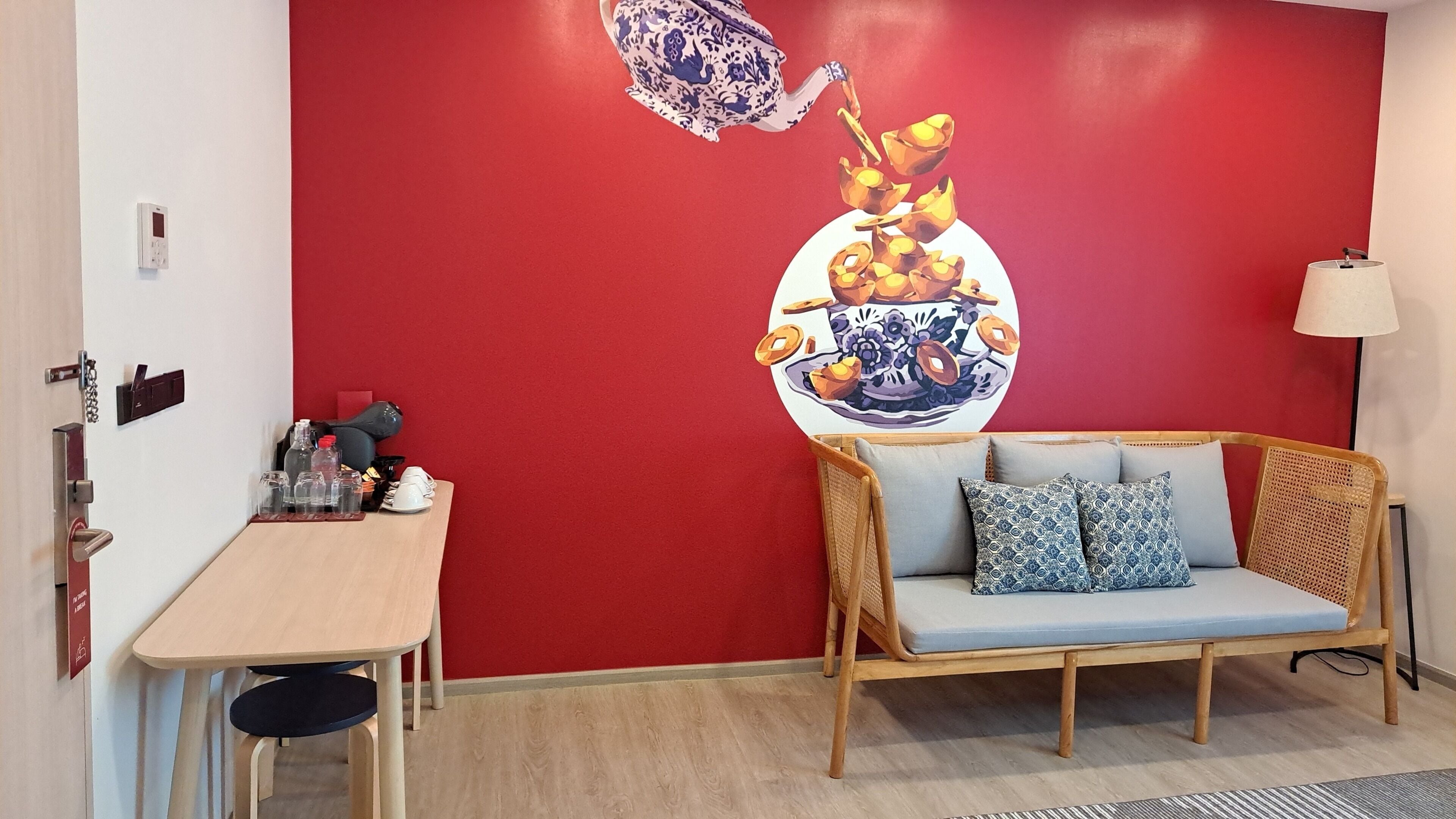Una habitación con una pared roja, una mesa de madera con varios objetos, un sofá de mimbre con cojines y una lámpara fijada a la pared.