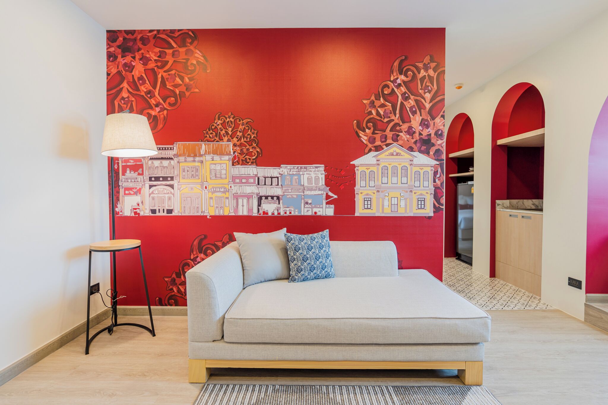 A modern hotel room with a large mural of colorful buildings, a sofa bed, a small table, and a lamp.