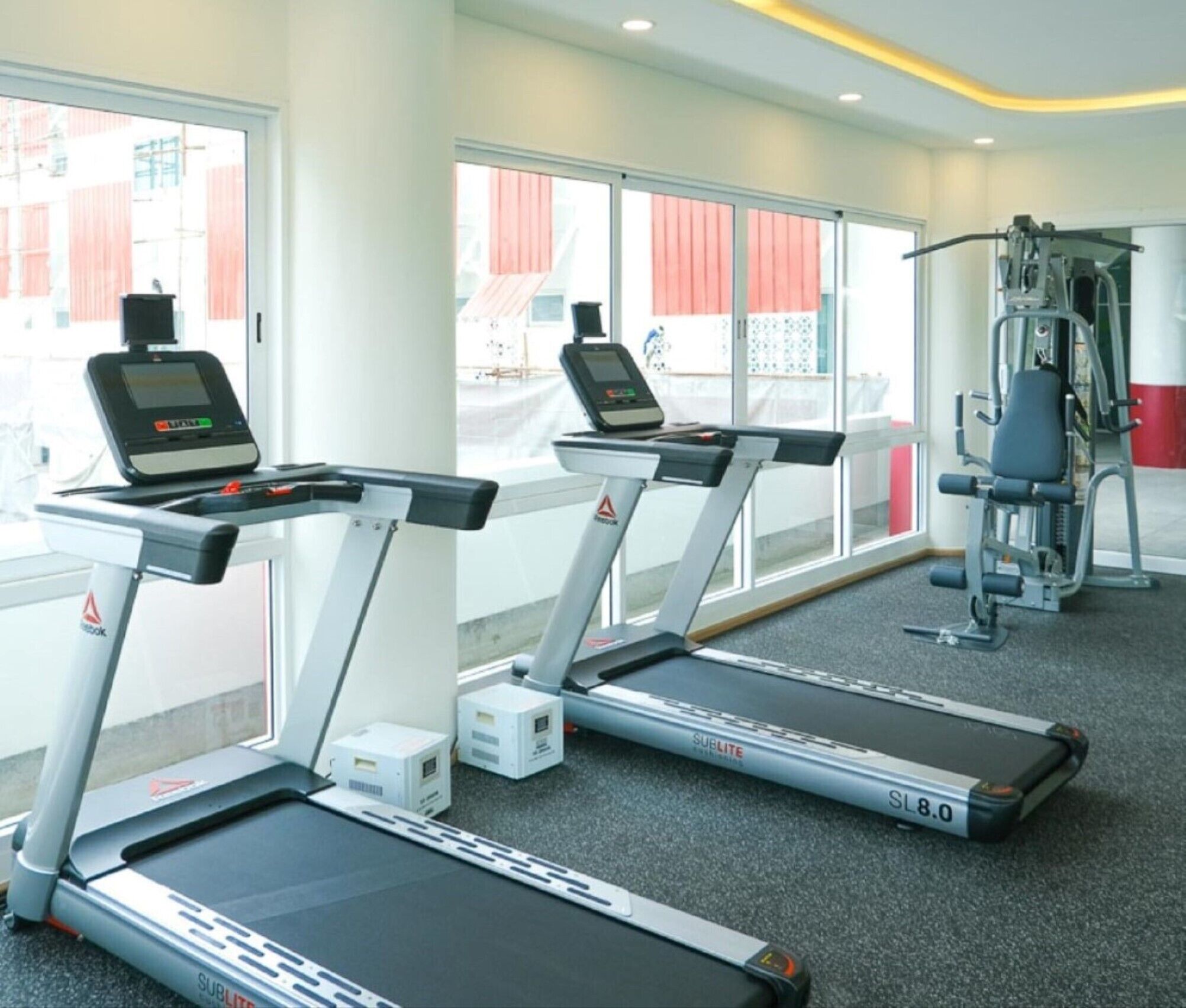 Une salle de sport équipée de tapis de course, d’appareils de musculation et d’une fenêtre donnant sur un bâtiment aux rayures rouges et blanches.