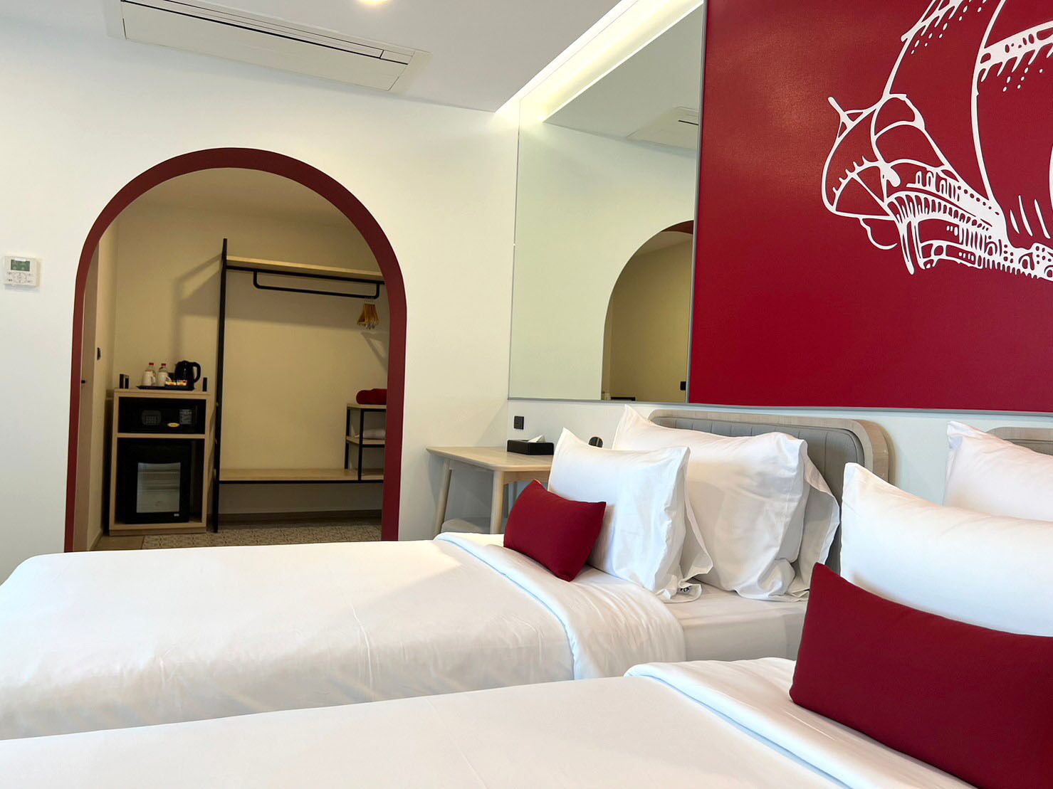 Habitación de hotel con dos camas, una pared de acento roja, un espejo grande y una kitchenette.
