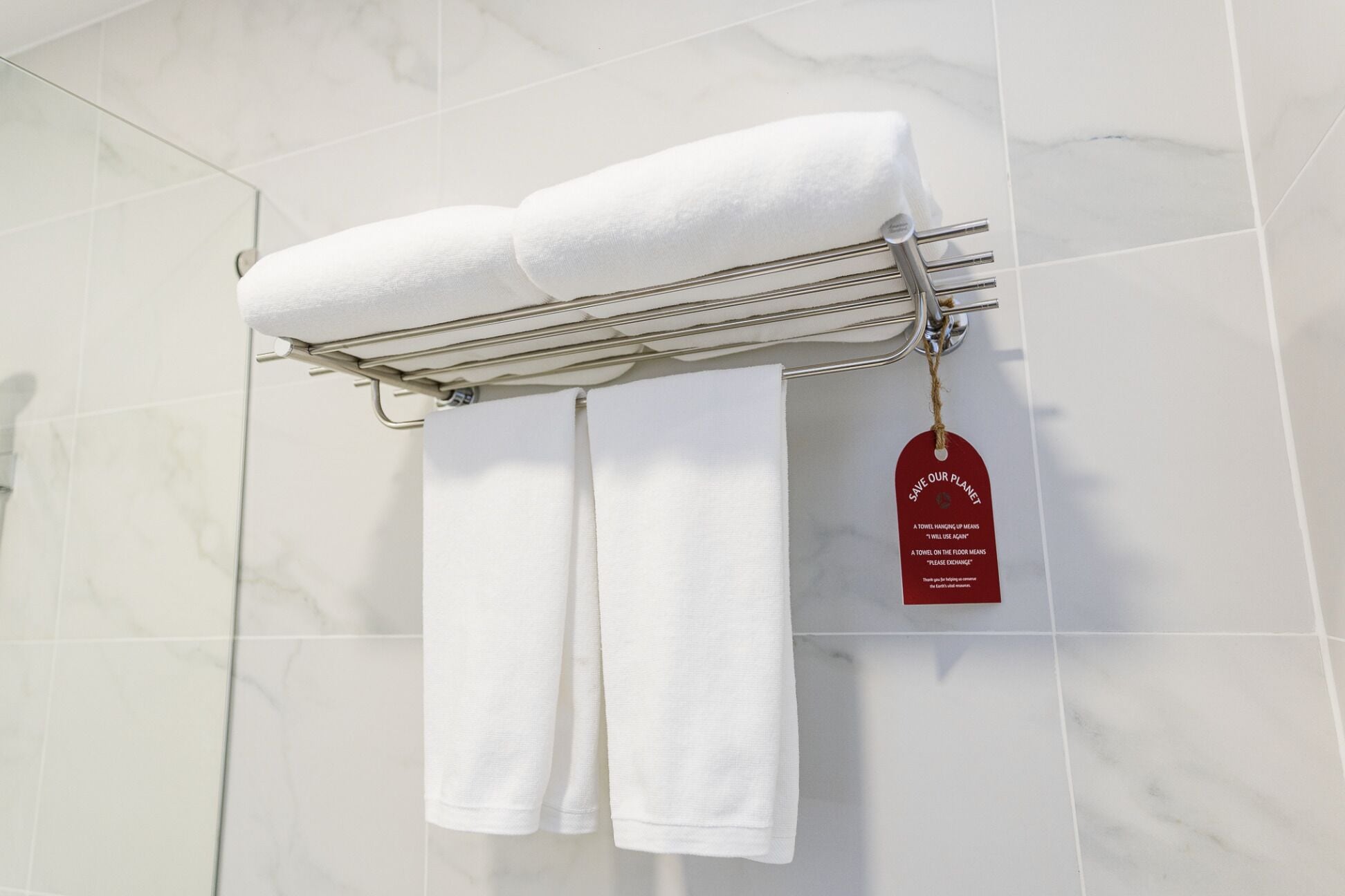A bathroom with white marble tiles, a towel rack holding two white towels, and a red tag with a message about towel usage.