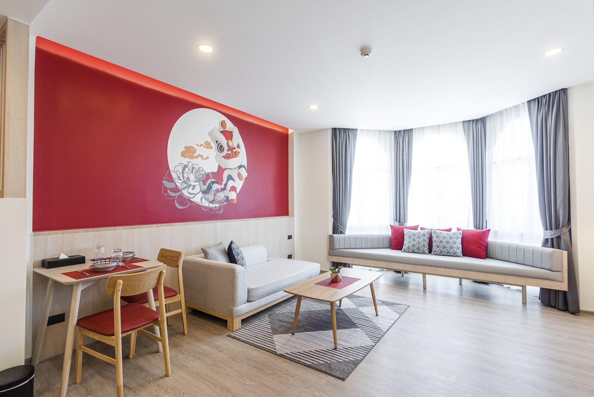 A modern living room with a red accent wall, a sofa, a dining table with chairs, and a large window with curtains.