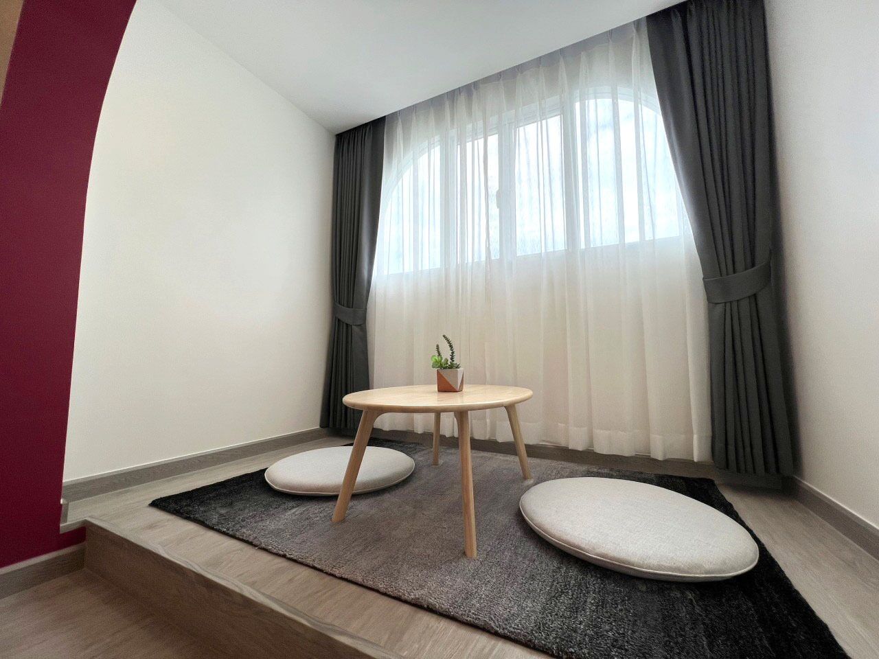 A small, modern room with a round wooden table, two cushioned seating areas, a small potted plant, and sheer curtains.