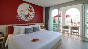 A hotel room with a large bed, a red wall with a traditional artwork, a balcony with outdoor seating, and a view of the city. - Hotel Midtown Ratsada (Phuket)