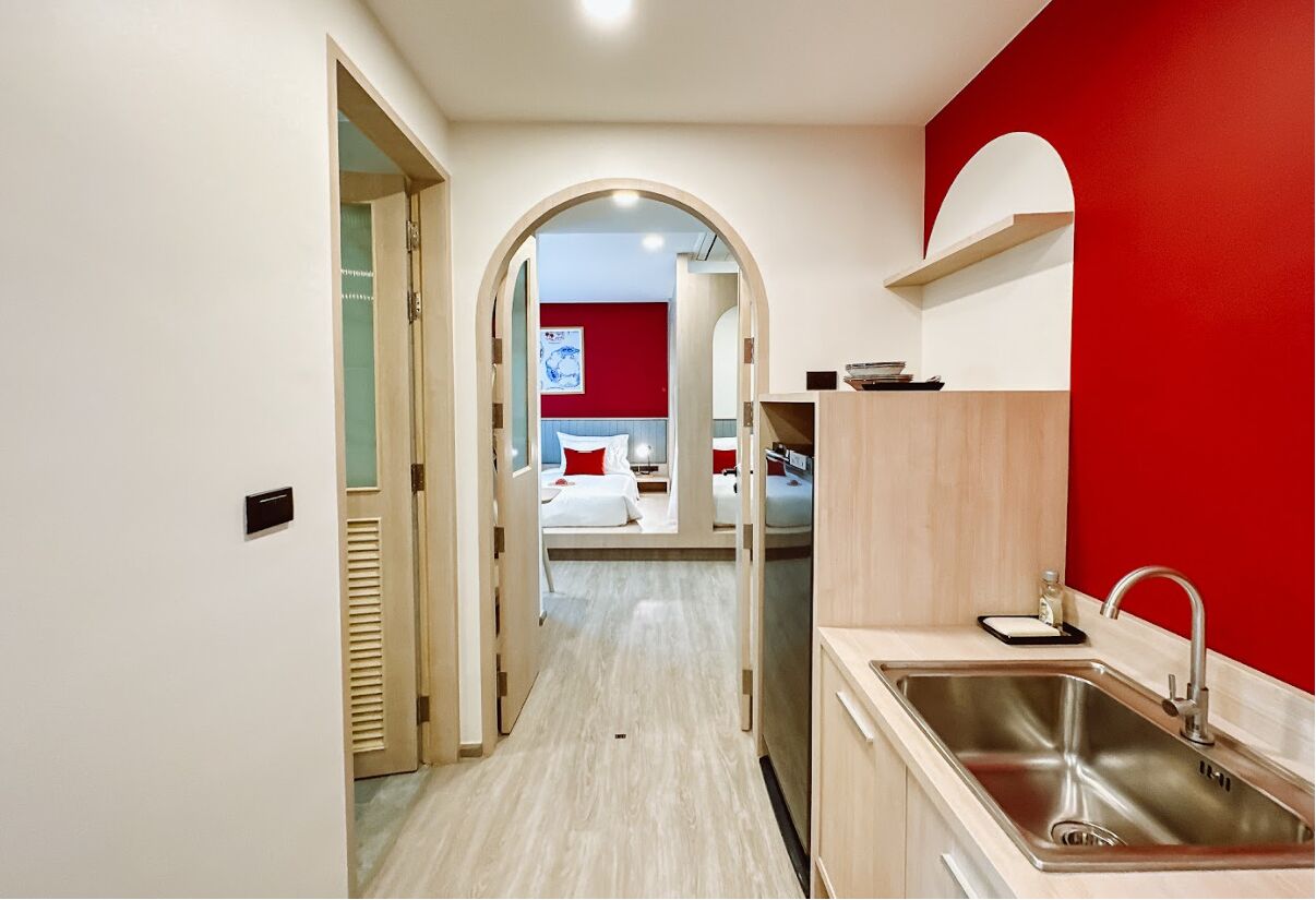 A hotel room with a kitchenette, a bed with red pillows, and a red accent wall.