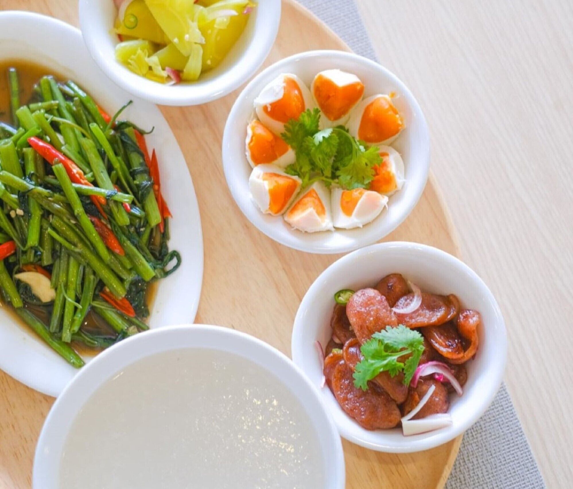 A wooden table with four bowls of food, including a dish with green vegetables, a bowl with orange slices, a bowl with meat and onions, and a bowl of clear soup.