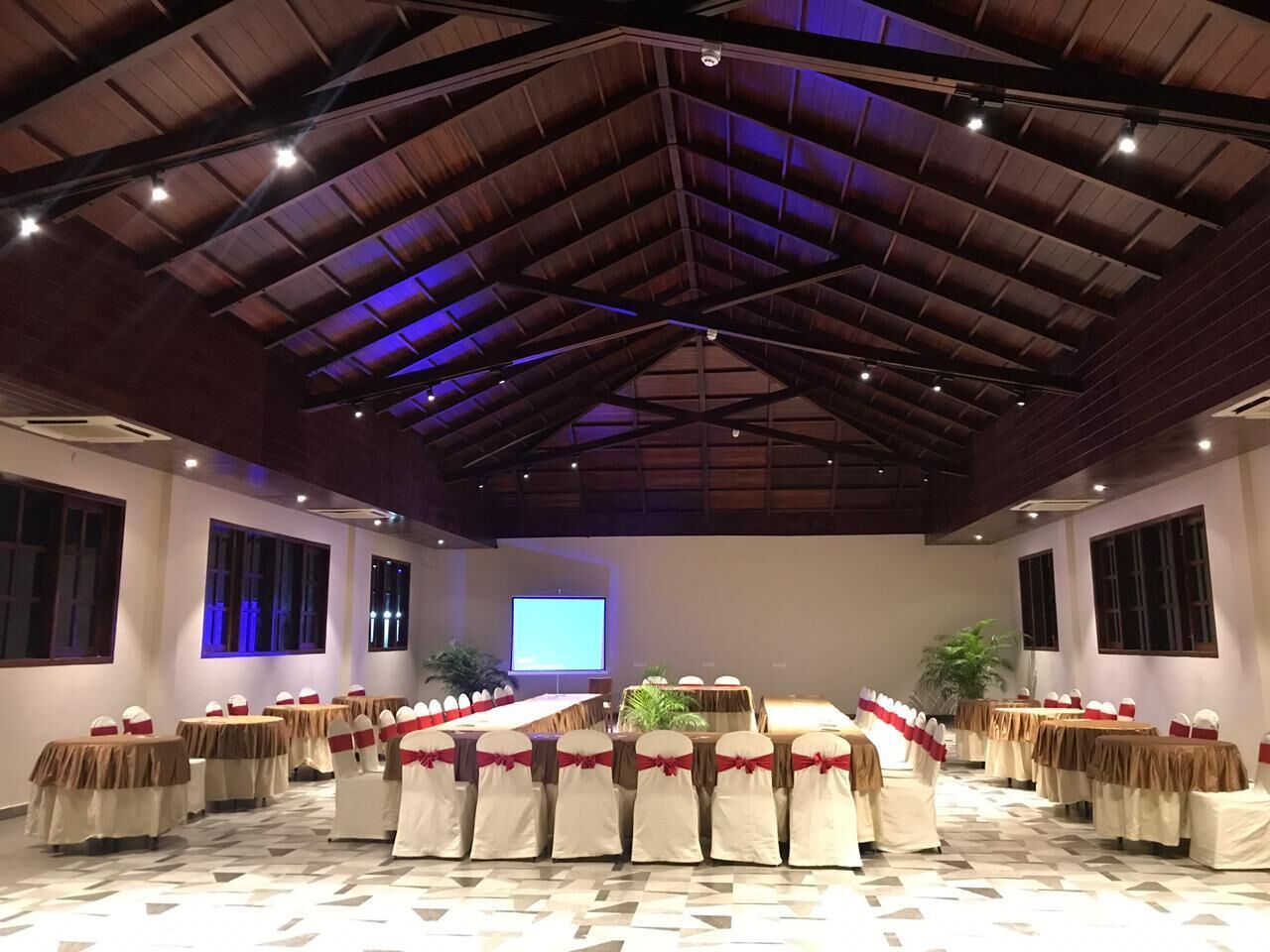 A conference room with a high ceiling, multiple tables with chairs, and a large screen at the front.