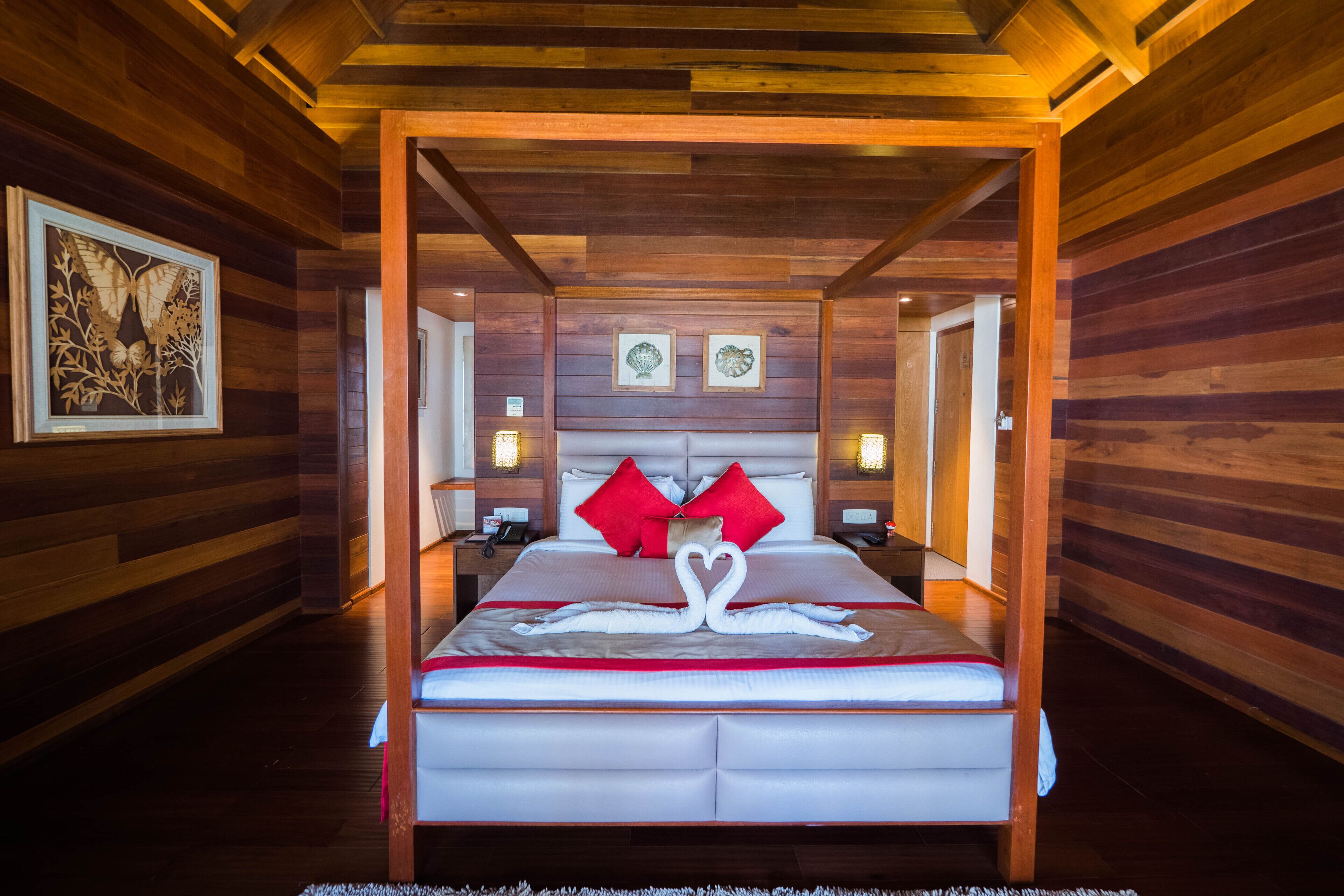 A wooden-paneled room with a bed featuring red pillows and a heart-shaped towel arrangement.