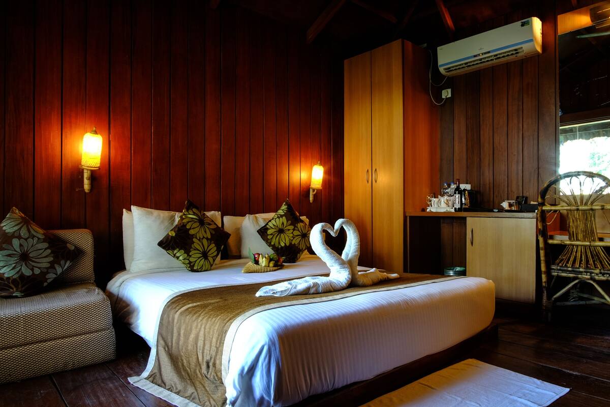 A hotel room with a large bed, two wall-mounted lamps, a wooden ceiling, a small kitchenette, and a wicker chair.
