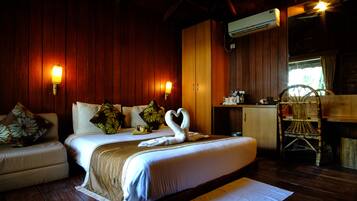 Habitación de hotel con una cama grande, dos lámparas fijadas a la pared, techo de madera, pequeña kitchenette y una silla de mimbre.