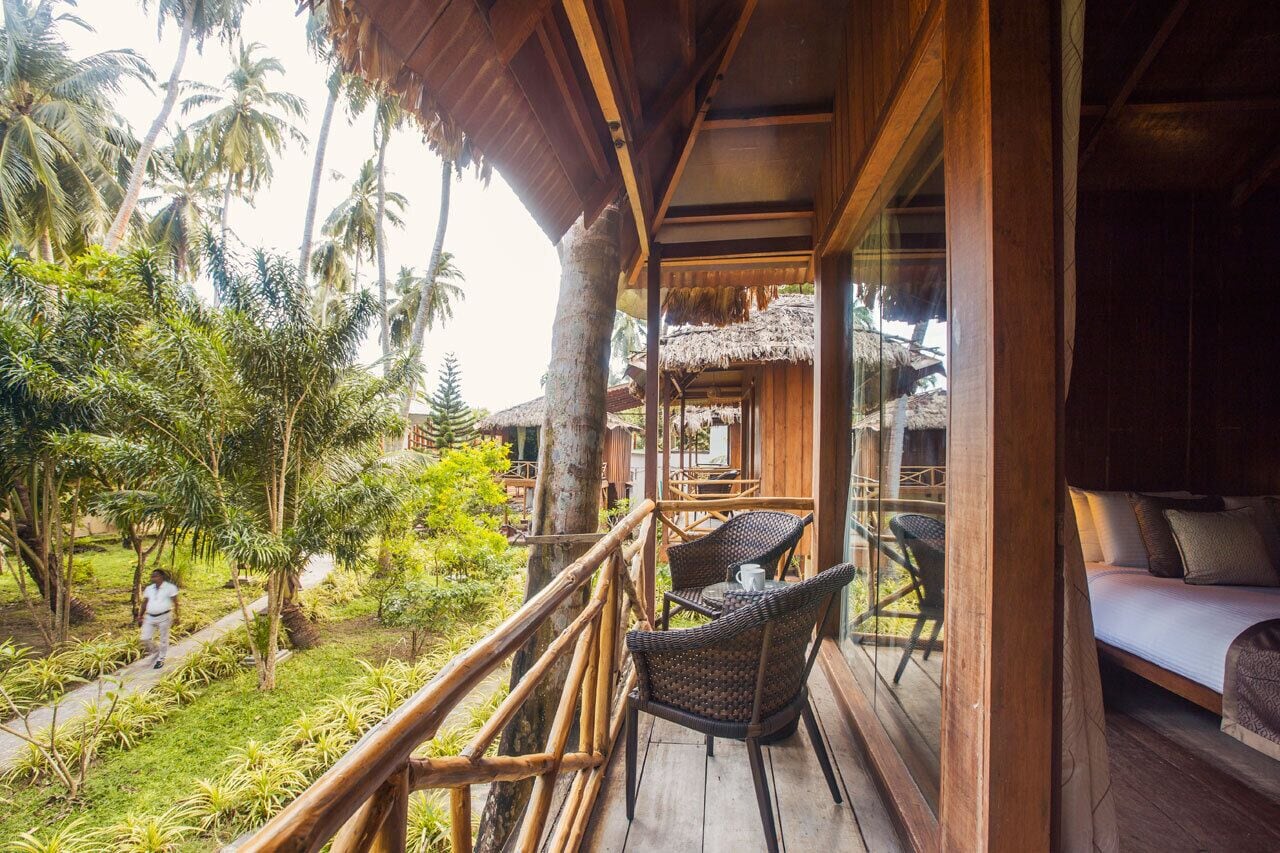 A wooden balcony with wicker chairs and a bed, overlooking a tropical garden with palm trees.