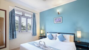 A hotel room with a large bed, two bedside tables with lamps, a window with curtains, and a wall-mounted picture. - Aastha Escape Resort (Calangute)