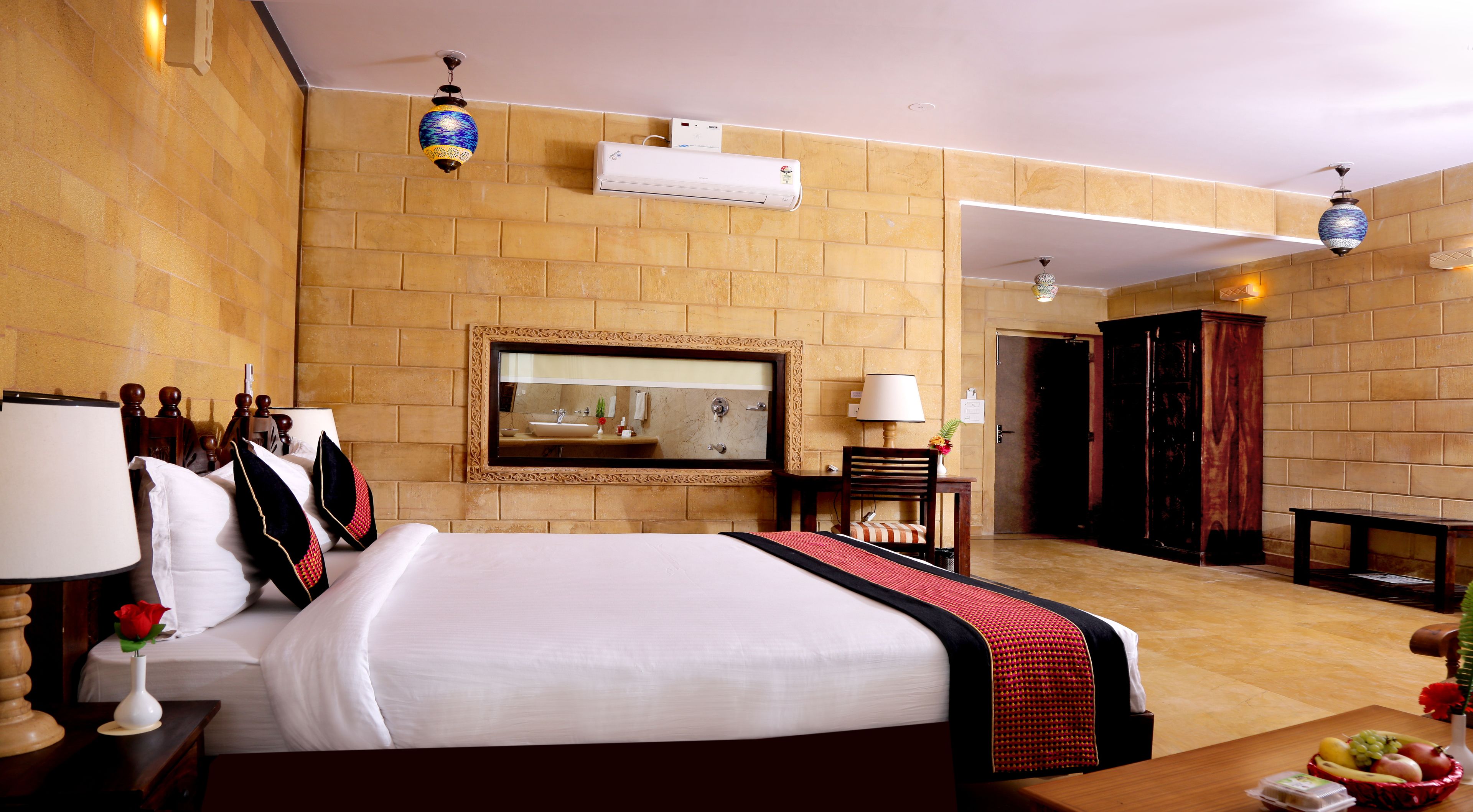 A hotel room with a large bed, a bathroom mirror, a bedside table with a lamp, a wooden chair, and a small table with fruit and a vase.