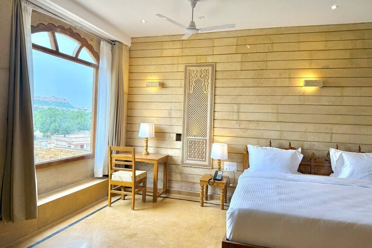 A hotel room with a bed, a desk, a chair, a ceiling fan, and a view of a mountainous landscape.