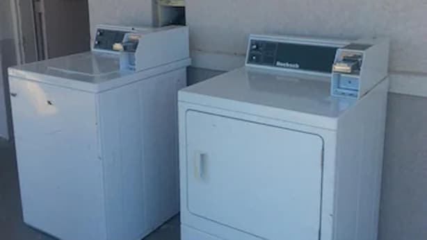 Two white front-loading washing machines in a room with a plain wall and a door in the background. - Budget Inn Fort Stockton (Fort Stockton)