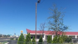 A commercial building with a red roof, a Shell gas station sign, and a fenced parking area with small trees. - Budget Inn Fort Stockton (Fort Stockton)