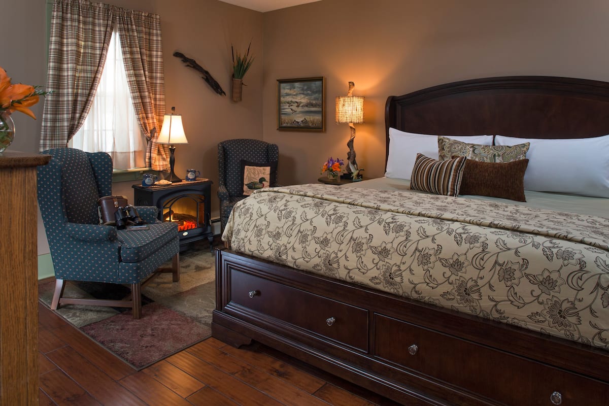 A bedroom with a large bed, a chair, a small table, a lamp, and a fireplace.