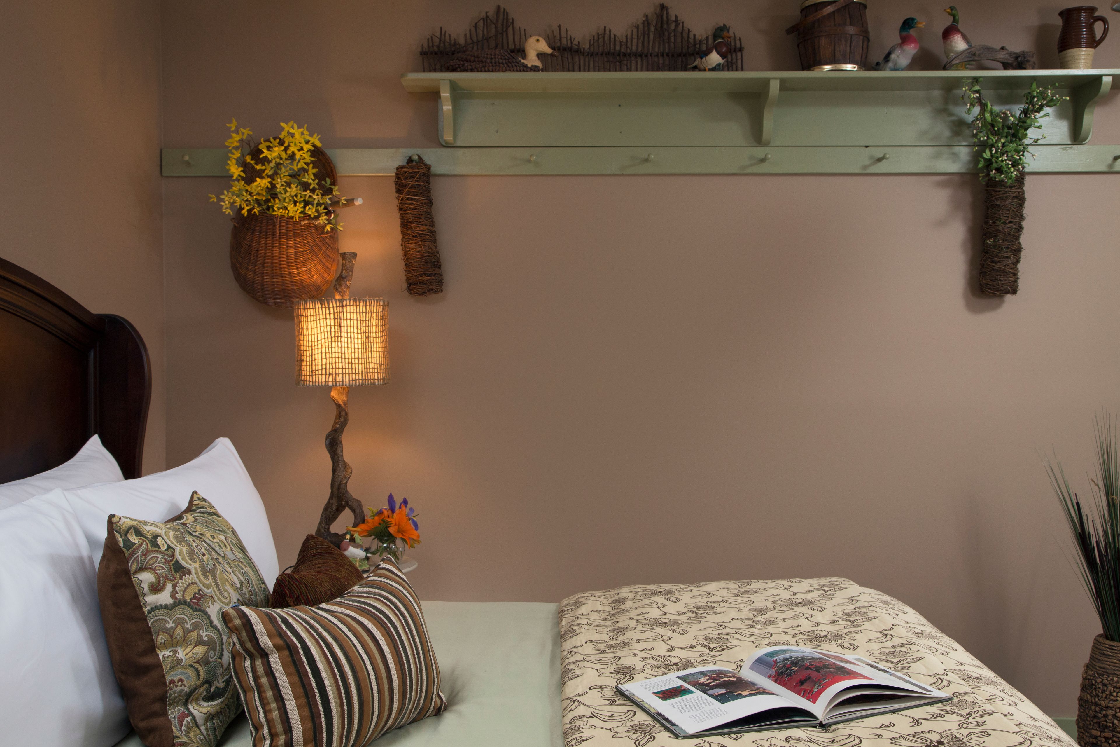 A bedroom with a bed, pillows, a bedside lamp, a shelf with decorative items, and a book on the bed.
