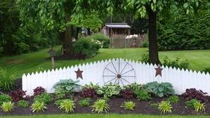 A garden with a white picket fence, various plants, and a wooden wheel. - 1825 Inn Bed and Breakfast (Palmyra)
