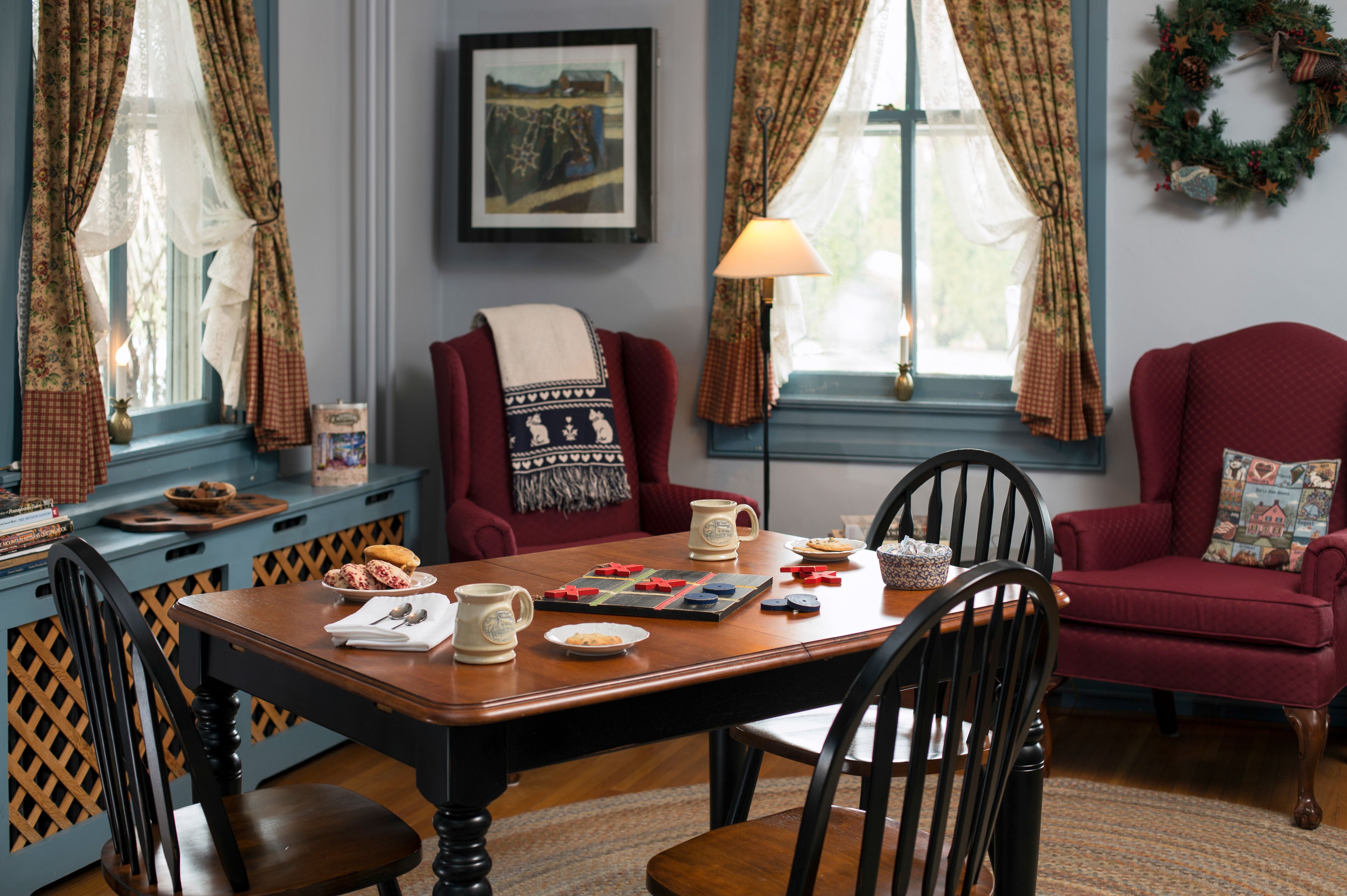 Un coin repas chaleureux avec une table en bois, des chaises et un fauteuil rouge. Une lampe et un tableau encadré décorent les murs.